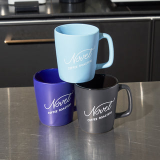 Blue coffee mug stacked on top of a purple and black coffee mug, all with Novel Coffee Roasters branding.