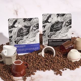 10 oz bags of rogue espresso and black swan espresso nestled among whole bean coffee beans and multiple espresso cups.