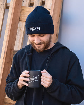 Man wearing black cuffed Novel beanie holding a black Novel Coffee Roasters coffee mug